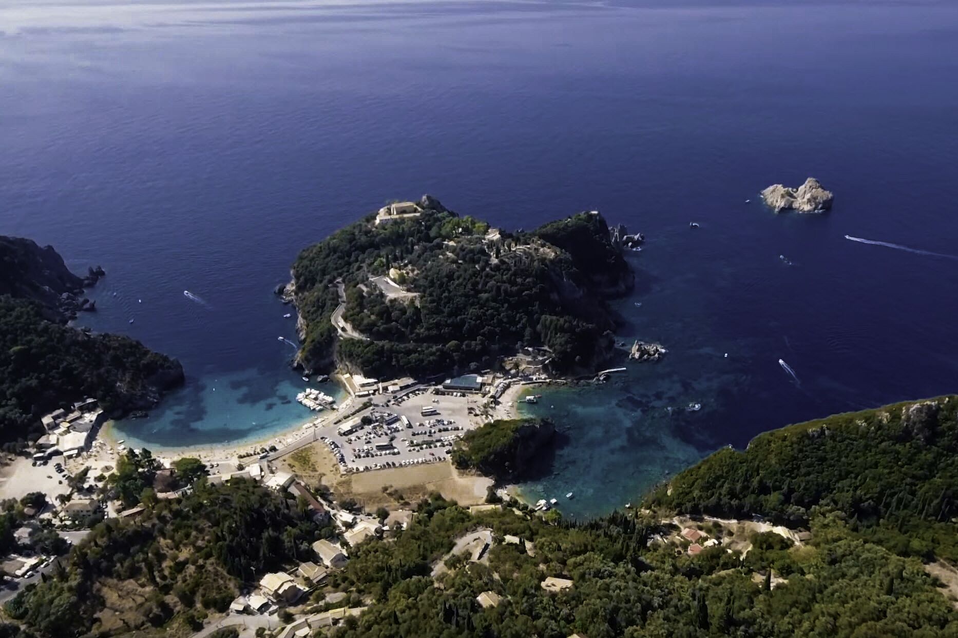 Vista aérea de una zona costera con una pequeña isla que cuenta con una playa, aparcamiento y vegetación densa.