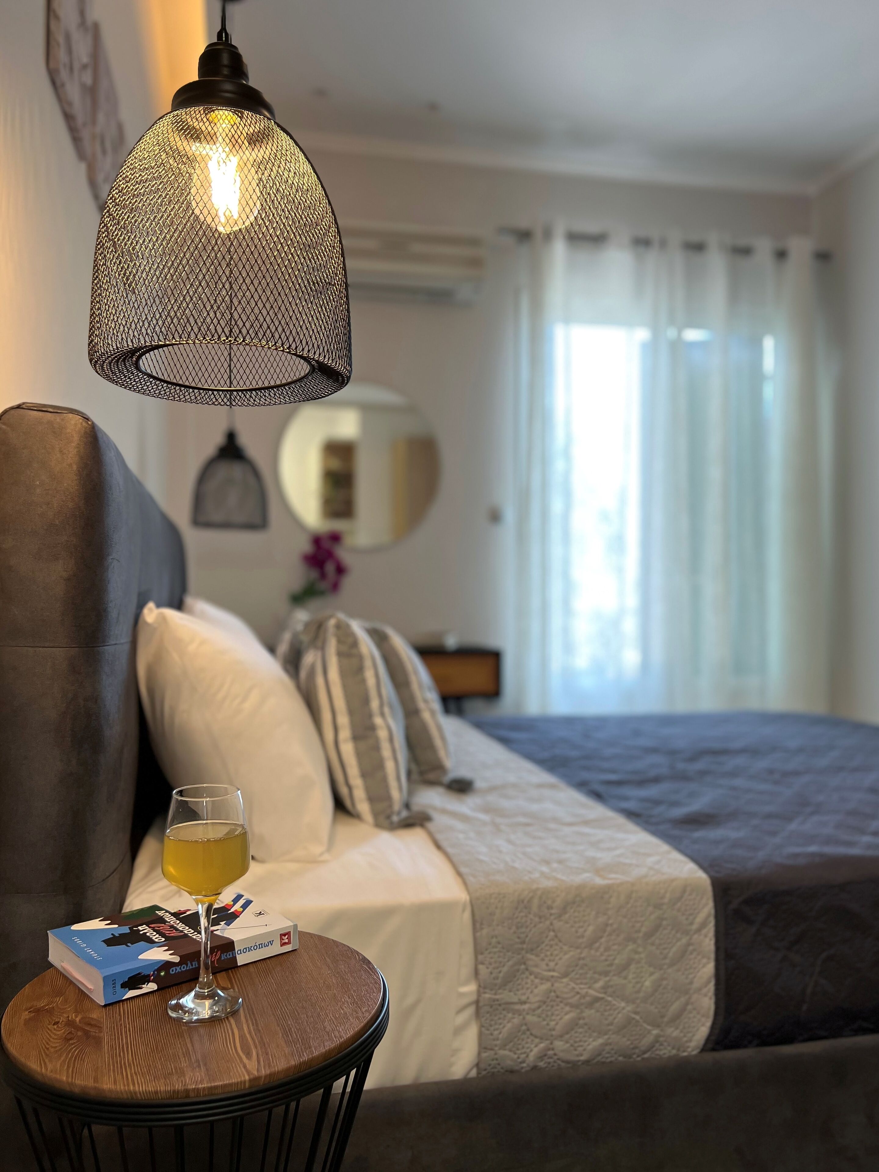Une chambre d’hôtel avec un lit, une table ronde sur laquelle se trouvent un verre et des livres, un luminaire suspendu et une fenêtre avec des rideaux.