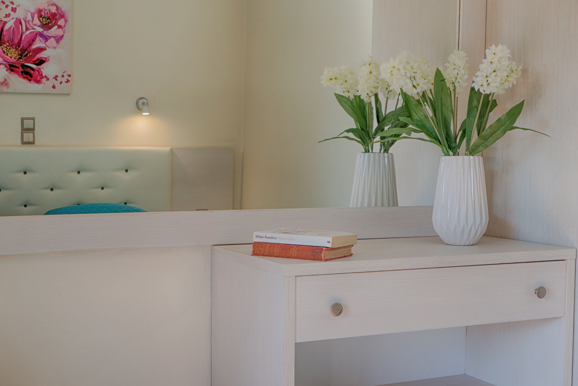 A white vanity with a mirror, a small white vase with white flowers, and a book titled "Milan Kundera Immortals".
