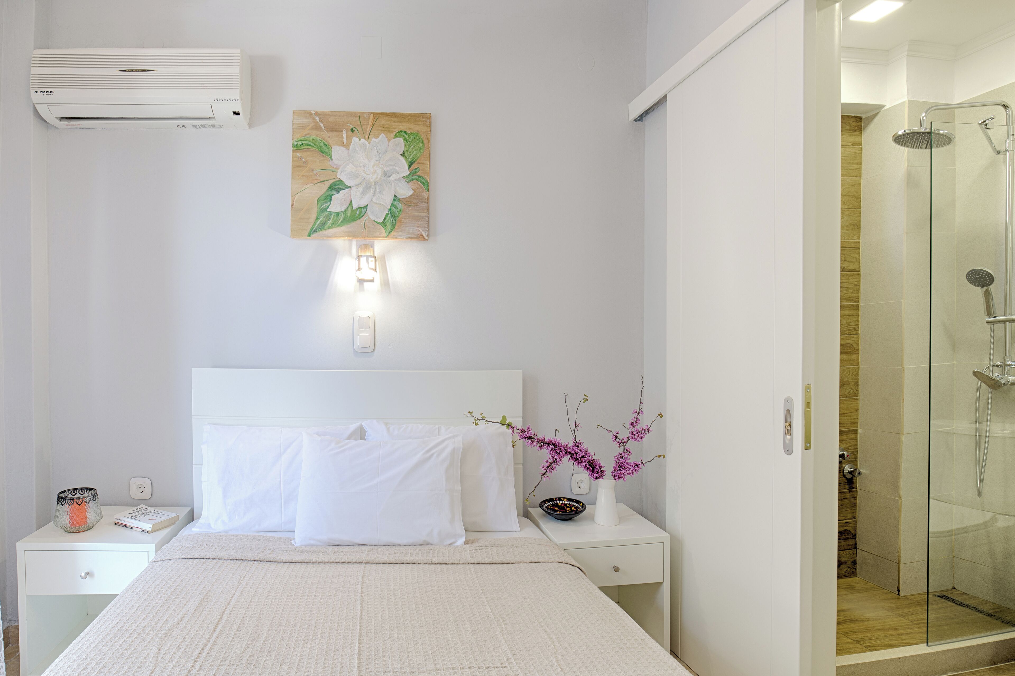 Une chambre d’hôtel comprenant un lit, une table de chevet, une commode, un climatiseur fixé au mur et une cabine de douche en verre.