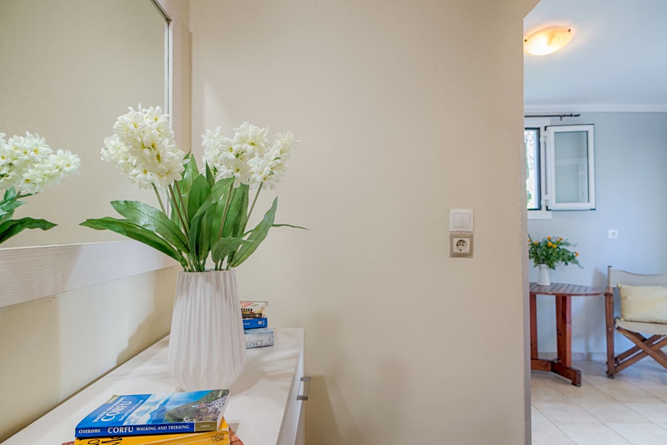 Un couloir avec un vase blanc rempli de fleurs blanches, posé sur une étagère blanche, une petite table avec des livres et, au fond, une chaise en bois.