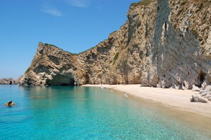 A coastal beach with clear turquoise waters, rocky cliffs, and a sandy shoreline.