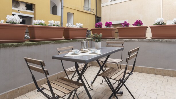 Eine Terrasse im Freien mit einem Tisch für zwei Personen, umgeben von Holzstühlen und Blumentöpfen mit Blumen.