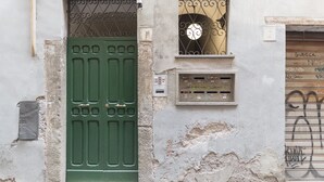 Eine verwitterte Gebäudefassade mit einer grünen Tür, einem kleinen Fenster mit dekorativem Metallgitter und einem Verteilerkasten.