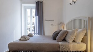 Cama bem arrumada com cabeceira, janela com cortinas e luminária fixada na parede.