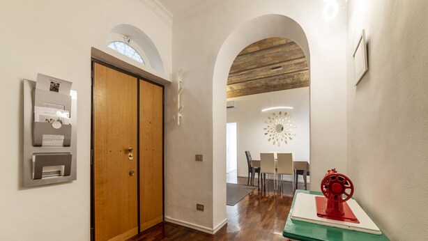 Um corredor com piso de madeira, uma porta de madeira, uma caixa de correio na parede e um objeto vermelho e branco sobre uma mesa.