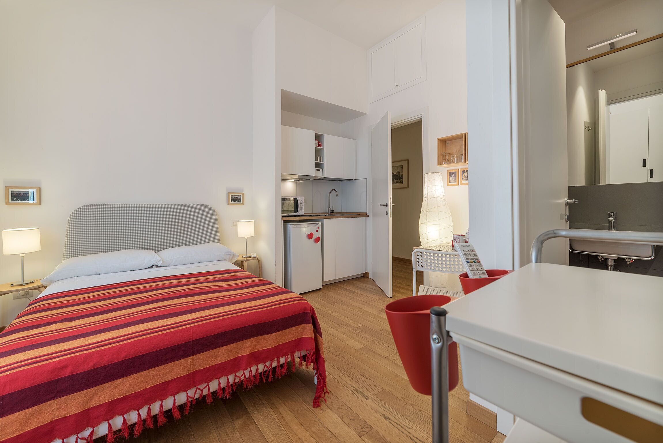 A bedroom with a bed, bedside tables, a red blanket, a kitchenette, and a bathroom with a mirror.