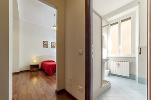 Quarto de hotel com piso de madeira, cama com cobertura vermelha, mesa de cabeceira com abajur, radiador fixado na parede e janela com vidro jateado.