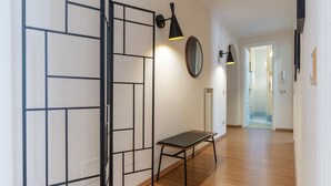 Un pasillo con piso de madera, una partición de metal negro y un banco. Hay dos lámparas de pared y un espejo.