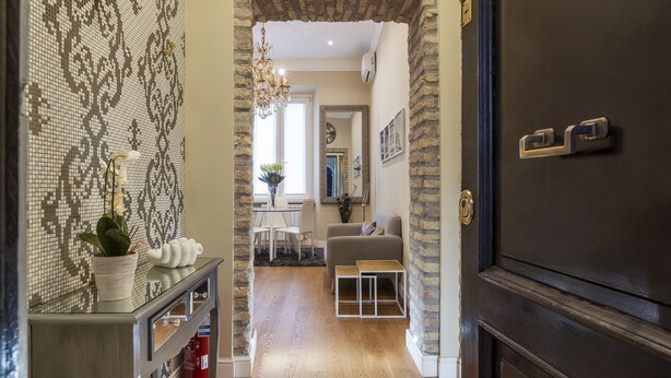 Un pasillo con una pared de mosaicos, suelo de madera, una puerta oscura y una zona de estar con una silla y una mesa.