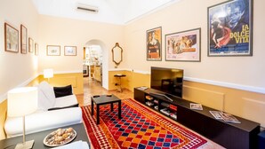 A living room with a sofa, TV, and a patterned rug. There are framed pictures on the wall and a poster of a movie.