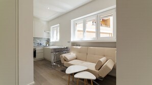A modern kitchen with white cabinets, a marble backsplash, and a dining area with a beige sofa and two white stools.