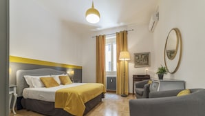 Quarto de hotel com cama, escrivaninha, cadeira, abajur, espelho e janela com cortinas.