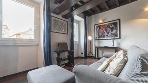 A living room with a sofa, a small table, and a lamp. There is a window with curtains, a painting on the wall, and a view of buildings outside.