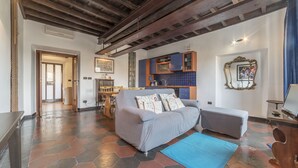 A living room with a sofa, a small table, and a blue rug. There is a kitchen area with cabinets and a sink. The room has a tiled floor and wooden beams on the ceiling.