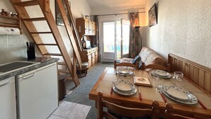 Uma cozinha compacta com escada de madeira, área de jantar com mesa posta para quatro pessoas e um sofá ao fundo.