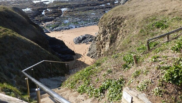 Uma trilha costeira que leva a uma praia com terreno rochoso e uma cerca de madeira.