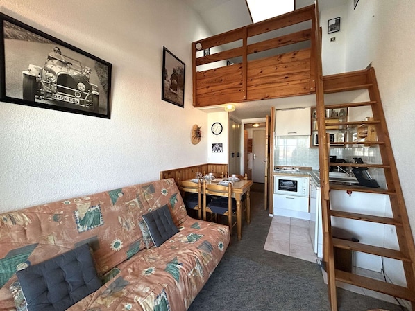 A cozy room with a wooden loft, a sofa with a patterned cover, a dining table, and a kitchen area with a stove and shelves.