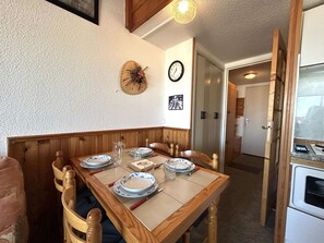 Ruokailutilassa on puinen pöytä, johon on katettu neljälle, seinäkello ja seinälle kiinnitetty koriste-esine.