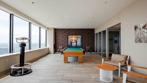 A modern room with a pool table, a telescope, and a large window offering a city view. - 18th FL Stylish CozySuites w/ roof pool, gym #1 (Dallas)