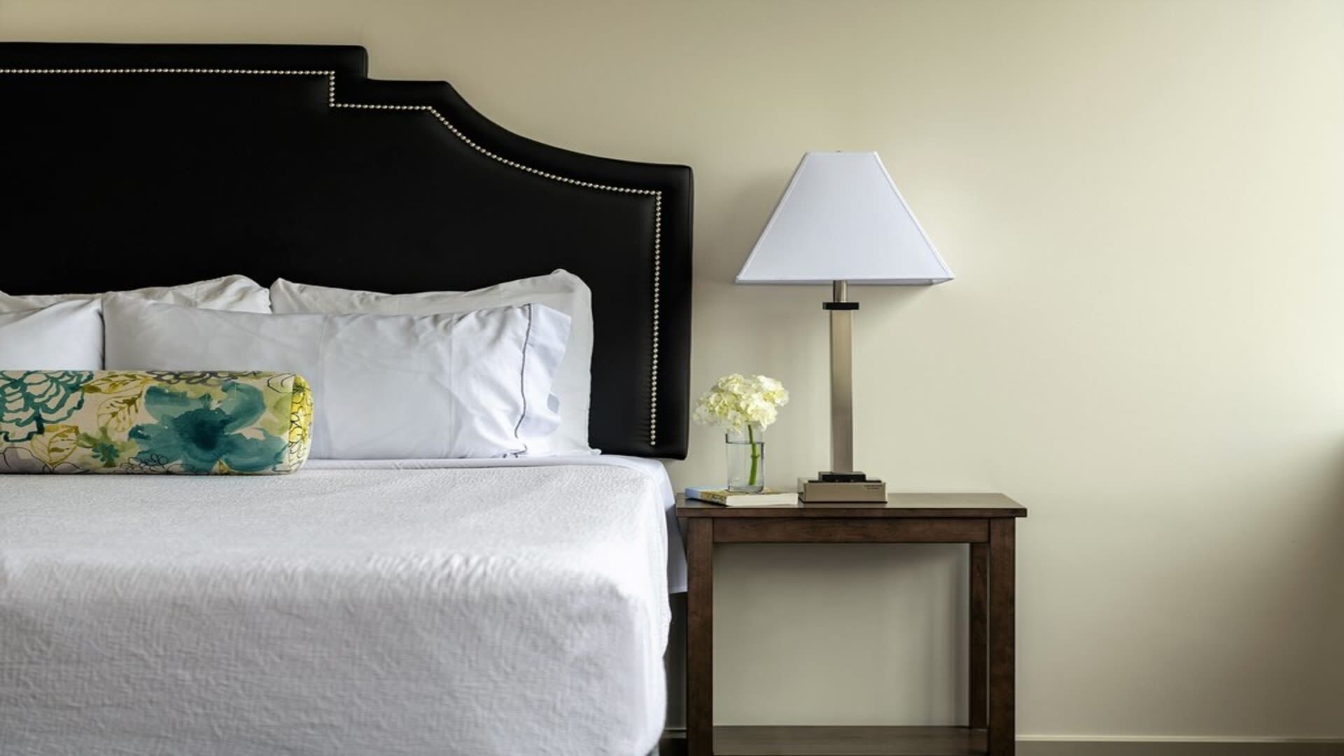 A neatly made bed with white linens and decorative pillows, a bedside table with a lamp and a vase of flowers, and a plain wall in the background.