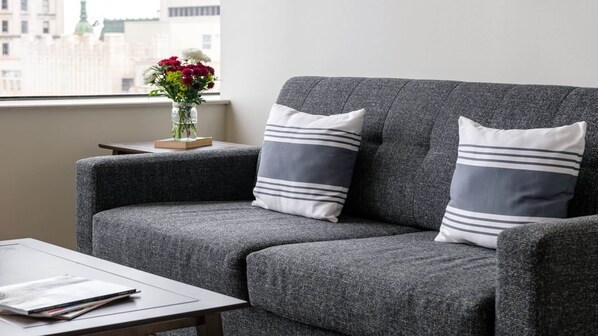 A grey sofa with striped pillows, a small table with a magazine, and a vase with flowers on a windowsill.