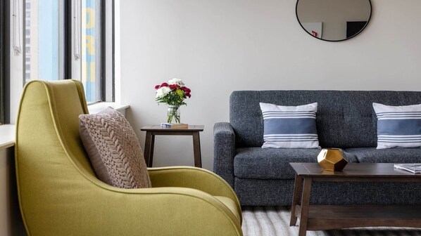 A modern living room with a green armchair, a grey sofa with striped pillows, a small table with a vase of flowers, and a round mirror on the wall. - 16th FL Bold CozySuites w/pool, gym, roof #1 (Dallas)