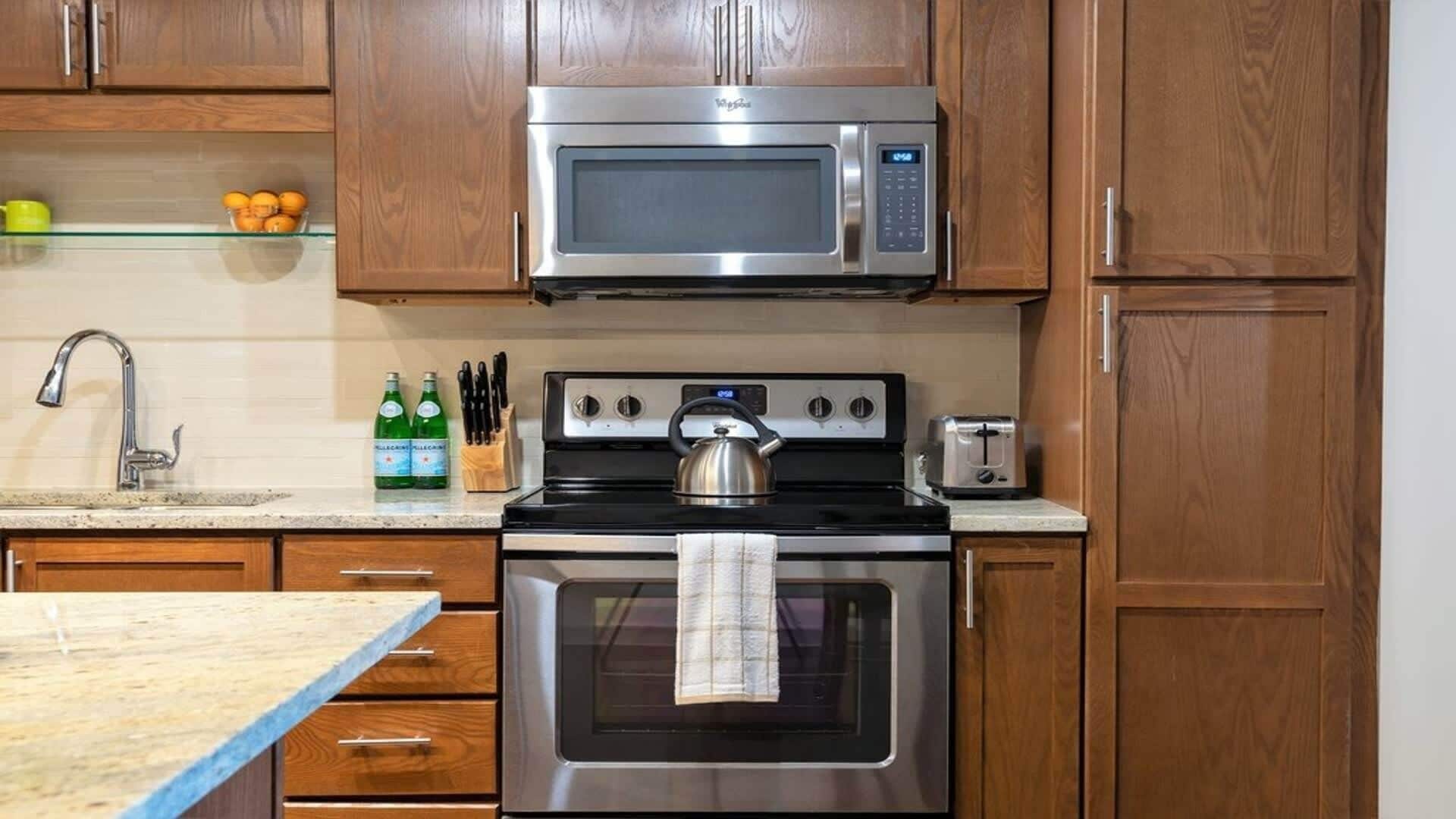 Una cocina con gabinetes de madera, un microondas de acero inoxidable, una estufa con un hervidor, un tostador y un lavamanos con grifo.