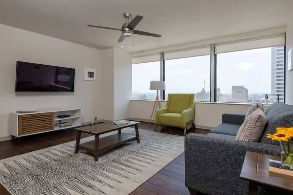 A modern living room with a flat-screen TV, a ceiling fan, a sofa, a coffee table, and a view of the city through large windows.