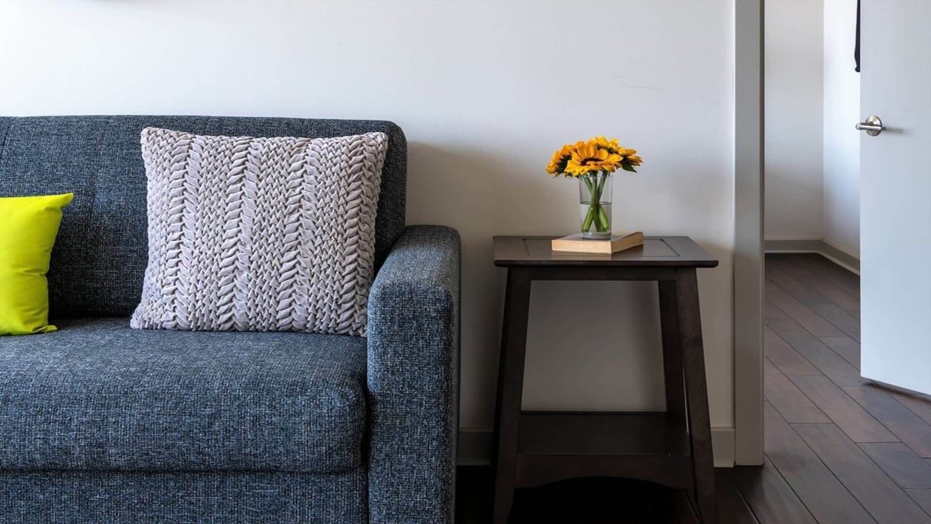 A grey sofa with a woven throw pillow and a bright yellow accent pillow. A small wooden side table with a vase of yellow flowers is placed beside the sofa.