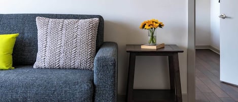 A grey sofa with a woven throw pillow and a bright yellow accent pillow. A small wooden side table with a vase of yellow flowers is placed beside the sofa.