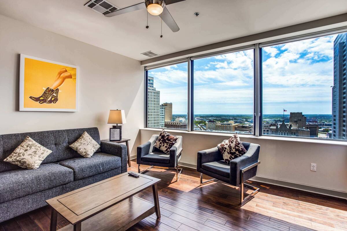 A modern living room with a grey sofa, two armchairs, a coffee table, and a large window offering a city view.