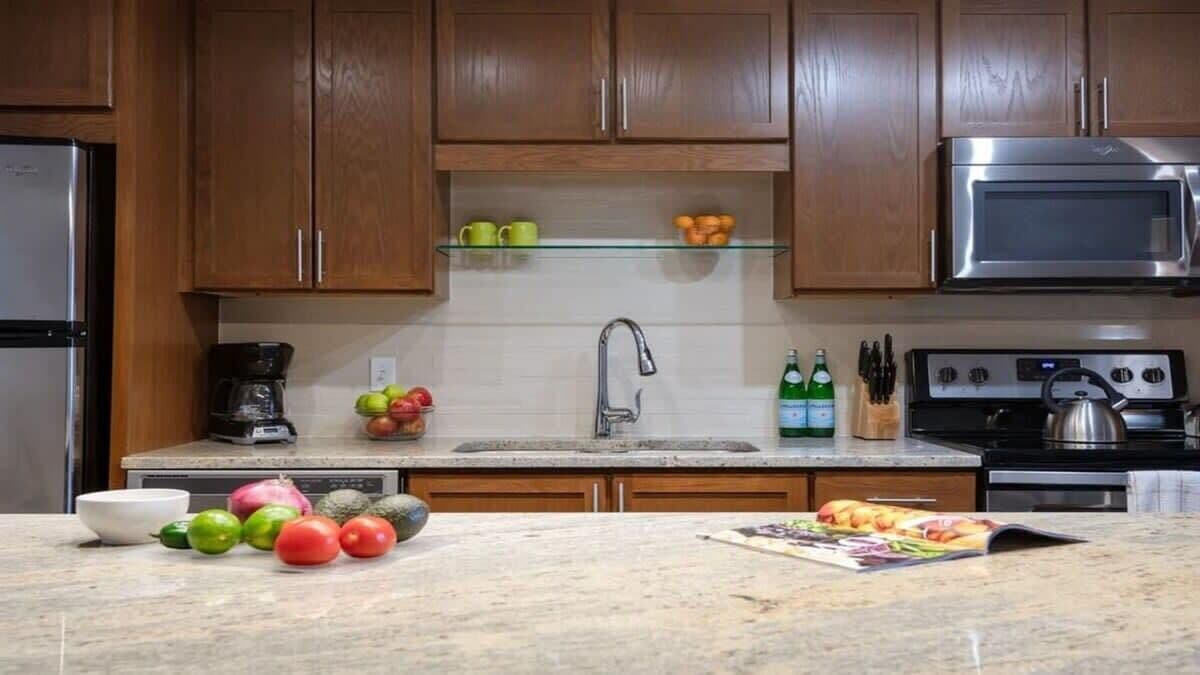 A kitchen with wooden cabinets, a stainless steel refrigerator, a microwave, a sink, and a countertop with fruits and a magazine.