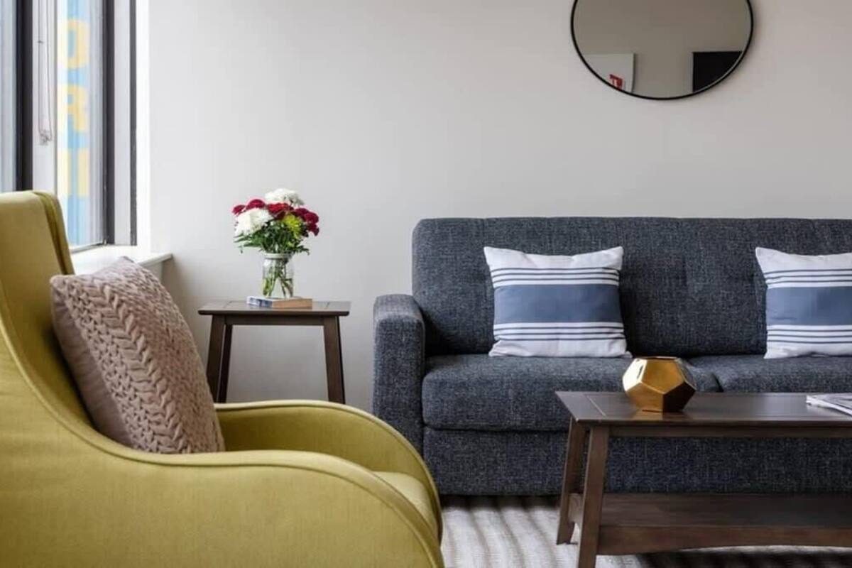 A living room with a yellow armchair, a grey sofa with striped pillows, a small table with a vase of flowers, and a round mirror on the wall.