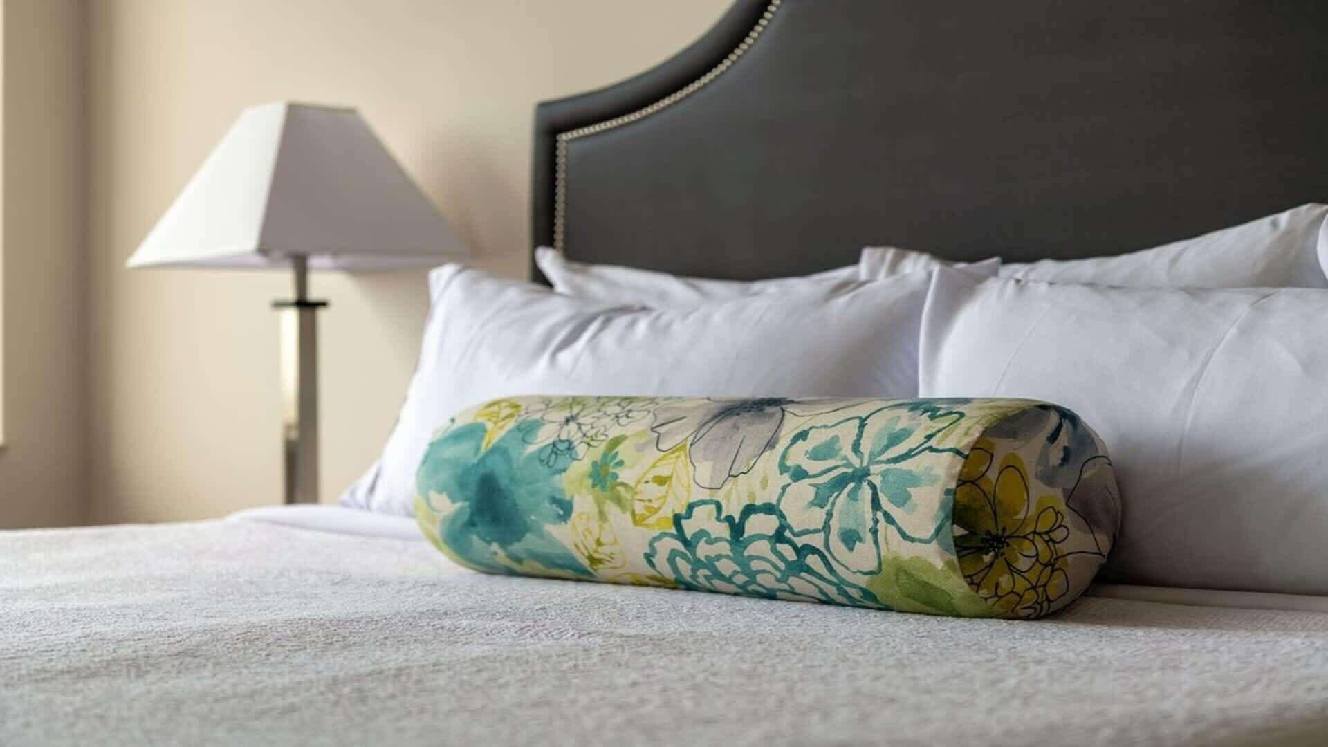 Cama bem arrumada com lençóis brancos, travesseiro decorativo com padrão floral e cabeceira escura.