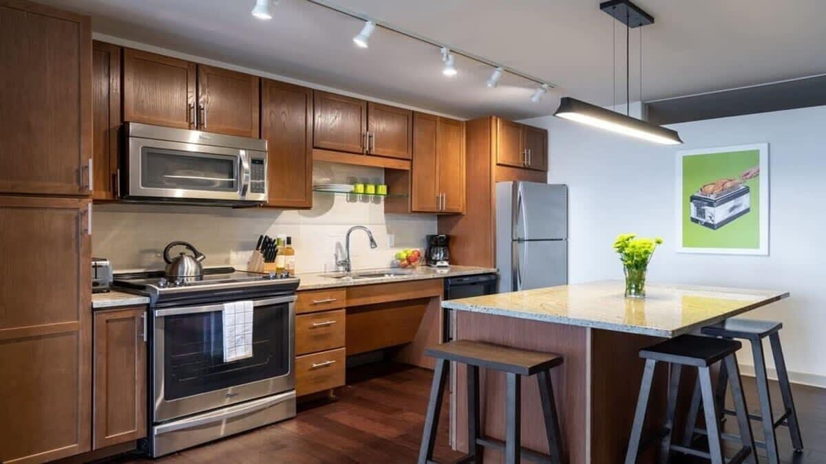 A modern kitchen with wooden cabinets, stainless steel appliances, a central island with stools, and a hanging light fixture.