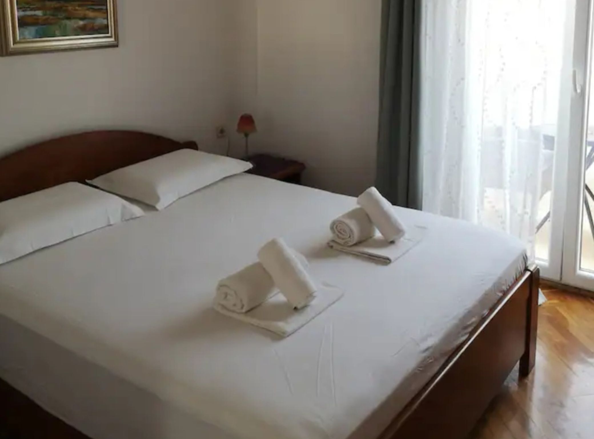 Een hotelkamer met een groot bed, witte lakens, twee opgerolde handdoeken, een nachtkastje, een lamp en een schilderij aan de muur.