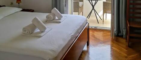 Una habitación de hotel con una cama bien tendida, una pequeña mesa con una silla afuera y piso de madera.