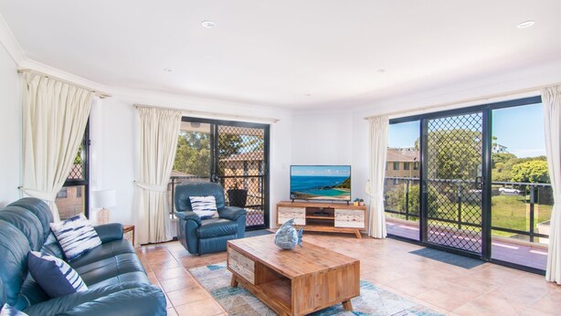 A spacious living room with a leather sofa, a wooden coffee table, and a TV on a wooden stand. There are large sliding glass doors leading to a balcony with a view of the sea. - Horizons 8 on the lake (forster)