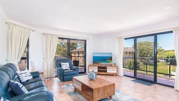 A spacious living room with a leather sofa, a wooden coffee table, and a TV on a wooden stand. There are large sliding glass doors leading to a balcony with a view of the sea.