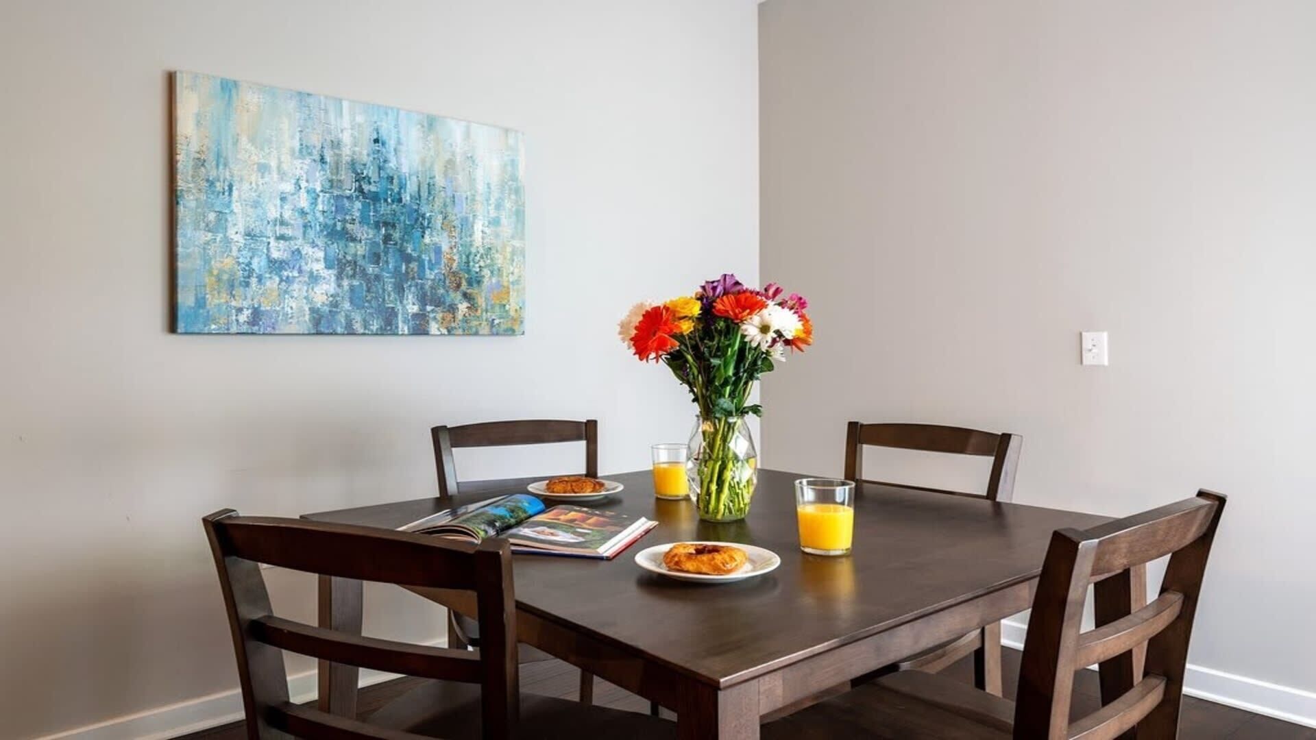 A dining table with four chairs, a vase with flowers, a plate with food, a glass of orange juice, and a magazine.