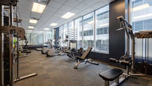 Un gimnasio con diversas máquinas de ejercicio, ventanas amplias y vistas a edificios.