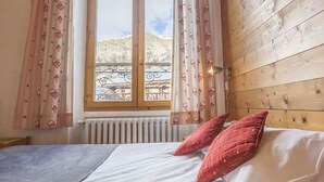 A bedroom with a wooden headboard, a bed with white linens and two red pillows, a window with a view of a balcony and mountains, and a radiator below the window.