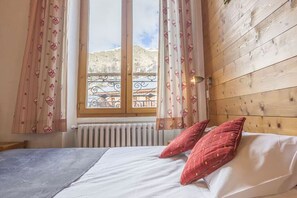 Quarto com cabeceira de madeira, cama com lençóis brancos e duas almofadas vermelhas, janela com vista para uma varanda e montanhas, e um radiador abaixo da janela.