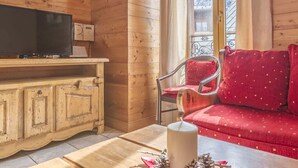 A cozy wooden cabin interior with a television on a wooden cabinet, a red sofa with patterned cushions, and a window with a view of snow-covered trees.