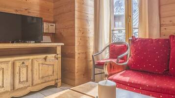 L’intérieur d’un chalet en bois chaleureux et accueillant, avec un téléviseur posé sur un meuble en bois, un canapé rouge agrémenté de coussins à motifs et une fenêtre donnant sur des arbres couverts de neige.