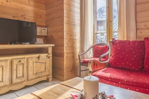Interior acolhedor de uma cabana de madeira com televisão em um armário de madeira, um sofá vermelho com almofadas estampadas e uma janela com vista para árvores cobertas de neve.
