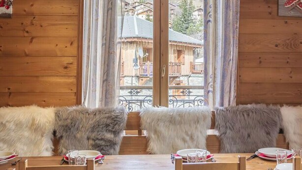 A wooden cabin with a dining table set for two, fur-covered benches, and a view of a mountain village through large windows.