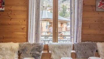 Une cabane en bois avec une table à manger dressée pour deux personnes, des bancs recouverts de fourrure et une vue sur un village de montagne, visible à travers de grandes fenêtres.