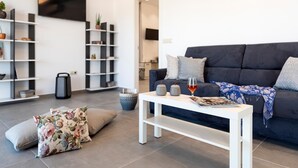 Un salon moderne avec un canapé foncé, une table basse blanche et des coussins à motifs floraux. On y trouve des étagères ornées d’objets décoratifs et un téléviseur à écran plat fixé au mur.
